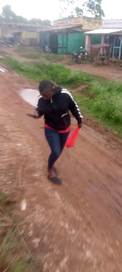 The image shows a young woman walking down a dirt road in Kivaywa, Kenya. She is wearing a black hoodie with white stripes, blue jeans, and red sneakers. She is holding a red cloth in her right hand. The road is muddy and there are puddles of water on the ground. The environment is rural and there are buildings in the background. The woman is walking in the direction of a shop on the right side of the image.