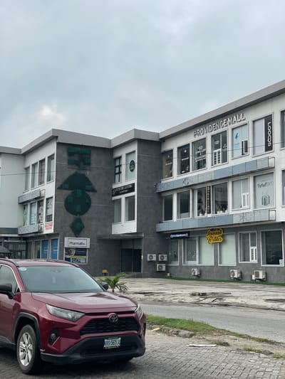 The image captures an exterior view of Providence Mall in Ilado, Nigeria. The mall is a multi-story building with a modern facade, featuring a combination of white and dark grey panels. The name "PROVIDENCE MALL" is prominently displayed in large, silver lettering across the top of the building. Various shop windows are visible, some with signage indicating businesses such as "JEWELLERY & ACCESSORIES", "Firstpa", "LOOQS", "LUXURY LIVING by Roro Casa", and "Jamiah Textiles".
In the foreground, a dark red Toyota RAV4 SUV is parked on a paved surface. The car's license plate reads "KRD-565 JQ". To the left of the mall, a portion of another building with the sign "BUILDERS H..." is visible. The ground area in front of the mall is a mixture of paved surfaces and some areas of sparse grass. Several air conditioning units are mounted on the exterior walls of the mall. The sky is overcast, suggesting a cloudy day. There are no people or active interactions immediately discernible in the scene.