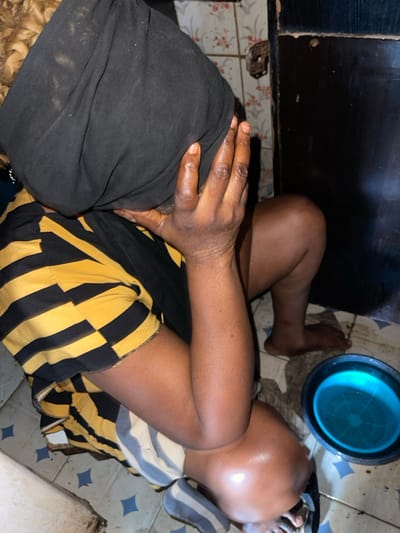 A person lies on a tiled floor in a cramped space. They wear a black head covering and yellow and black striped shorts. Their arms are crossed on their stomach. A blue basin sits beside their head, and a striped cushion supports their head. The lighting is dim.