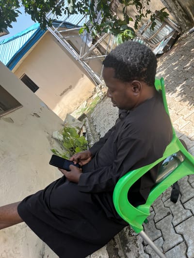 A person sits on a bright green plastic chair on a paved outdoor area. The individual, facing to the right and looking down at a smartphone, is dressed in dark clothing. The smartphone is held in both hands. The environment includes a building with a pale wall and a blue roof, and lush green foliage. The ground is covered with interlocking concrete pavers. The image appears to be taken in an outdoor setting within Galadimawa Village, Nigeria.