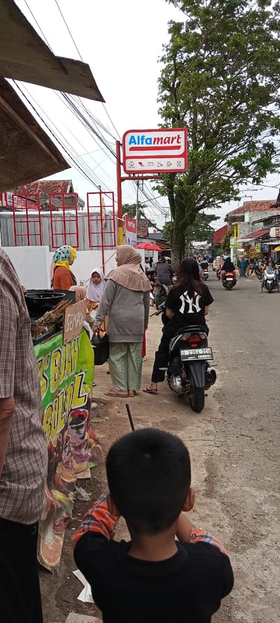The image shows a scene in Cangkuang, Indonesia (latitude: -7.056, longitude: 107.553) of several people in front of an "Alfamart" store. A man wearing a plaid shirt stands on the left side of the image, with a large sign with the words "Buah Bulu" behind him. There are four women, one of whom is wearing a white headscarf and a grey jacket, another wearing a black headscarf and an orange shirt, another is wearing a white headscarf and an orange shirt, and the last wearing a black shirt with the letters "NY" printed on the back. The woman with the "NY" shirt sits on a black motorbike. The man and the woman with the "NY" shirt are the only people whose faces are visible in the image. On the right side of the image is a view of a street with parked and moving motorbikes. There are several other people in the background, but their faces are not visible. A young boy with black hair stands facing away from the camera wearing a red, white, and blue plaid shirt over a black shirt. A large red sign reading "Alfamart" with some symbols and the words "2313 000" is visible. The image appears to be taken from a public place.