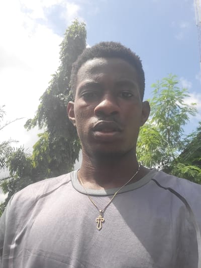 A medium shot, selfie-style photograph features a young Black male from the chest up, facing the camera. He has short, dark, curly hair and a fair complexion. He is wearing a grey, short-sleeved t-shirt with a subtle textured pattern and a darker grey stripe on the shoulder. A thin gold chain with a cross pendant rests on his chest. The background consists of lush green trees and a bright blue sky with scattered white clouds. The image appears to have been taken outdoors, possibly in a tropical or subtropical region. The lighting suggests it is daytime. The overall impression is casual and informal. The location is identified as Egbagu, Nigeria.