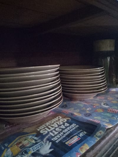 A close-up, slightly blurry, indoor shot shows stacked plates on a surface covered with a patterned cloth and some packaging. The plates are white with a dark rim and are arranged in two stacks. The stack on the left is closer to the camera and appears to be larger, with at least ten plates visible. The stack on the right is slightly further back and has fewer plates visible. Underneath the plates, a blue packaging material is visible with some text and images. The background is dark and appears to be the inside of a cabinet or shelf. The lighting is dim, creating shadows. There are no people or animals present in the image. The location is not discernible from the visual information provided.