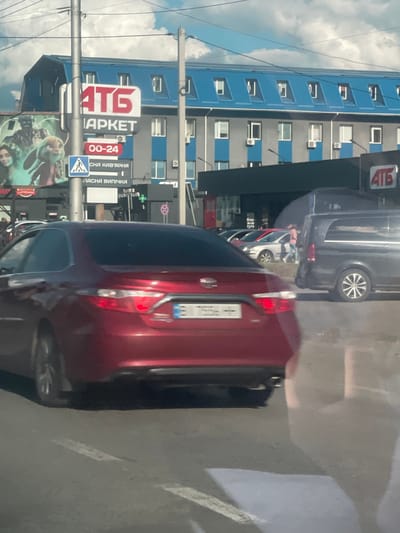 The image shows a street scene in Poltava, Ukraine, with a red sedan in the foreground. Behind the car, there is a large building with a blue roof and gray walls. A sign for "ATG Market" is visible on the building. To the left of the building, there is a billboard with images of characters. Several other cars and a van are parked or driving in the background. The sky is partly cloudy.