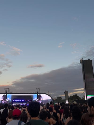 The image depicts an outdoor event, likely a concert, occurring at dusk in Martínez, Argentina. A dense aggregation of individuals, viewed from behind, occupies the foreground and midground, oriented towards a distant stage. The sky above transitions from a clear, light blue at the zenith to a lower expanse dominated by scattered clouds, some exhibiting faint pink and white illumination. The main stage structure in the background features multiple large video screens. These screens project identical close-up images of a female performer, characterized by dark hair and visible earrings. To the far right, a tall, dark, rectangular screen tower stands vertically. A line of trees is discernible on the horizon behind the stage. Numerous attendees in the immediate foreground and midground are holding up smartphones, many with their screens illuminated, indicating active recording or photography of the event.