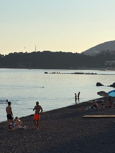 The image depicts a beach scene in Rafailovići, Montenegro, along the Adriatic coast. The scene captures people enjoying various activities. Along the beach, the sand slopes down to the water's edge. Several individuals are seen on the beach; some are in the water, swimming or wading, while others are on the shore, possibly resting or sunbathing. The individuals closer to the shoreline are clearly visible, showcasing their attire. Behind the beach, vegetation densely covers the mountainous terrain. Further into the sea, a string of buoys marks off a swimming area. In the distance, buildings and possibly more of the city are visible, adding depth to the view. The sky is light, suggesting either early morning or late afternoon.