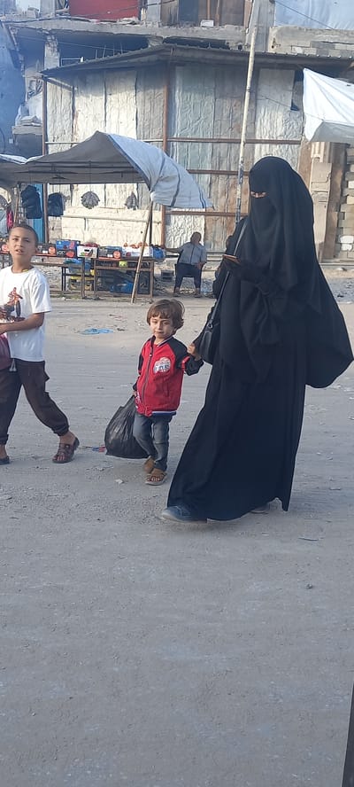 The image shows a street scene in Khan Yunis, Palestinian Territories. In the foreground, a young child wearing a red jacket and jeans walks with a black plastic bag in their left hand. To the left of the child, a boy in a white t-shirt and dark pants walks in the same direction. To the right of the child, a woman in a black niqab and abaya walks, holding a black phone in her gloved right hand. A black purse is slung over her left shoulder. In the background, there are market stalls with awnings and goods displayed. Further back, a man sits on a chair. The environment appears to be a somewhat dilapidated urban area with buildings showing signs of damage or disrepair. The ground is paved with dirt and gravel.
