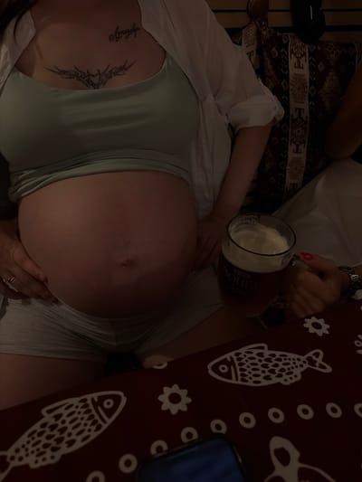 The image shows an overhead view of a pregnant person lying down, with their belly prominently displayed. They are wearing a light green tank top and a white open shirt. Tattoos are visible on their chest. A hand, adorned with a ring, is gently resting on their belly. In the background, a person is holding a large mug of beer, with foam spilling over the rim. The scene appears to be indoors, possibly in a casual setting like a bar or restaurant, as indicated by the presence of the beer mug and a patterned tablecloth with fish and flower motifs. The lighting is dim, suggesting it might be evening or a dimly lit establishment. The overall mood seems relaxed and intimate. The beer mug has visible text, which appears to be a brand name.
