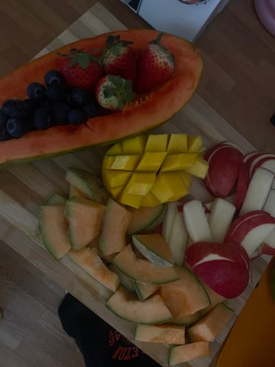 A wooden cutting board is arranged with a variety of fresh fruit, including a halved papaya filled with blueberries and strawberries, a mango cut into cubes, cantaloupe melon cut into wedges, and red and white apple slices. The board is situated on a wooden floor. The location is likely a home in Segovia, Spain, based on the provided coordinates. The image captures a still life, showcasing the different fruit and its preparation for consumption. The board is partially in shadow, indicating an indoor environment. There is no person visible in the image. The image suggests an activity of food preparation or consumption.