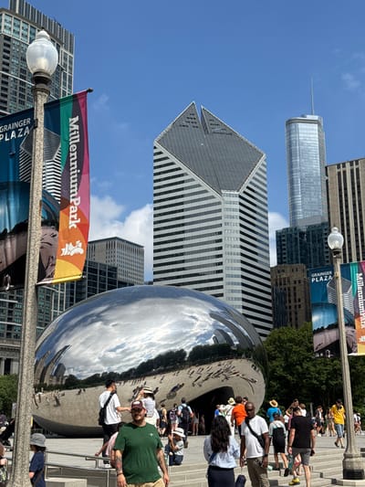 This media file captures a sunny day in Chicago's Millennium Park, featuring the iconic "Cloud Gate" sculpture, often referred to as "The Bean." The highly reflective surface of the sculpture mirrors the surrounding skyscrapers, the bright blue sky filled with scattered clouds, and the numerous people present. Several individuals are seen interacting with the sculpture, taking photos, and enjoying the park. Some are walking on the stairs and pathways around The Bean, while others are gathered near it. The scene suggests a tourist destination and a popular public gathering spot.
Notable details include the clear blue sky and bright sunlight, indicating it's likely midday. The architecture of the skyscrapers is prominent, reflecting in the sculpture, and some signage is visible. One banner reads "Millennium Park," and another smaller one beneath it says "GRAINGER PLAZA." A flag with colorful stripes, also bearing the "Millennium Park" text and a stylized figure, is attached to a lamppost. The mood appears lively and relaxed, typical of a pleasant day in a bustling urban park. The image is oriented vertically, with the sky and upper parts of the buildings appearing on the left and the ground and people on the right, as if the camera was tilted.