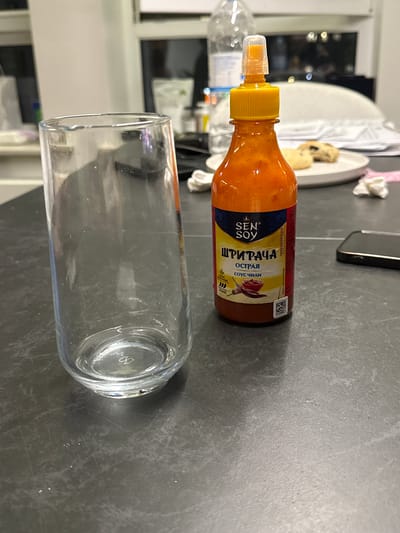 A close-up shot shows a bottle of Sen Soy Sriracha hot sauce placed horizontally next to a clear glass. The bottle is orange and red, with a yellow cap and a transparent nozzle. The label on the bottle features the Sen Soy logo and text in Russian, including "ШРИРАЧА," "ОСТРАЯ," and "соус чили." A small image of a chili pepper and a rose is also visible on the label. The glass is empty and transparent, with a thick base.

The scene appears to be indoors, likely on a kitchen counter or table, given the dark gray textured surface. In the background, out of focus, hints of what might be kitchen cabinets, lights, and a plate with food can be seen. The lighting suggests it might be evening or a dimly lit room. The overall mood is neutral, focusing on the presentation of the hot sauce and the glass. The image is captured with a shallow depth of field, blurring the background and highlighting the main subjects.