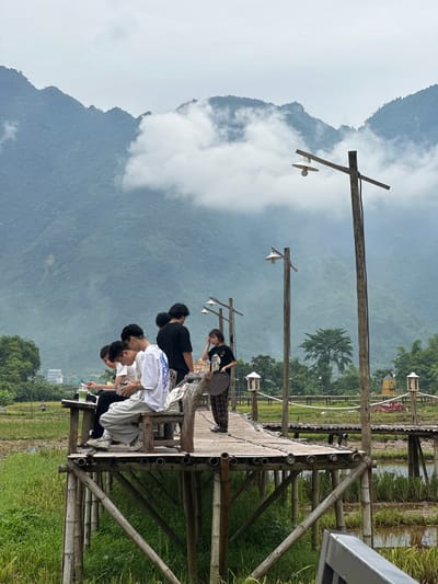 This image depicts a wooden boardwalk extending over a rice paddy in Mai Chau, Vietnam. Several individuals are present on the boardwalk, some seated and others standing. A group of young men are seated on a bench on the left side of the boardwalk, with one holding a mobile phone and another looking at his phone. A young woman stands on the boardwalk to the right, holding a round object and looking towards the left. The boardwalk is constructed from bamboo and wood, with wooden posts supporting it above the water and vegetation of the rice paddy. In the background, lush green mountains rise, partially obscured by white clouds. Several lampposts are visible along the boardwalk. The overall scene suggests a scenic viewpoint or a recreational area within a rural landscape.