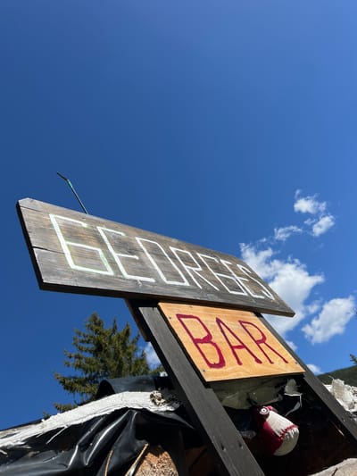 A weathered wooden sign with hand-painted letters is tilted against a dark wooden frame. The top plank displays the word "GEDPRIS" in white block letters outlined in green. Below it, a smaller wooden plank reads "BAR" in red capital letters. The sign is set against a bright blue sky with a few wispy clouds. In the background, a dark green evergreen tree is visible, along with a hint of a mountainous landscape. The lower part of the structure supporting the sign is covered in black plastic sheeting, with some torn white material peeking out. A small, stuffed object with red and white fabric and a pink button eye is partially visible beneath the "BAR" sign. The overall scene appears to be outdoors, possibly in a rural or mountainous area, with no identifiable urban features of Samokov, Bulgaria.