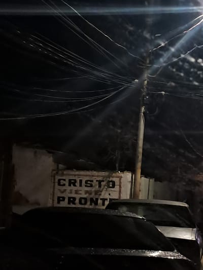 The image captures a nighttime scene in Cumana, Venezuela, with a prominent utility pole and a tangle of overhead wires dominating the upper portion. Below, a building with a weathered facade is visible, featuring a sign with the text "CRISTO VIENE PRONTO" (Christ is coming soon) in block letters. A portion of a vehicle's roof and windshield is visible in the foreground, suggesting its presence on a street or in a parking area. The overall lighting is dim, with artificial light sources creating lens flares and illuminating parts of the scene. The environment appears to be an urban setting, characterized by the presence of electrical infrastructure and buildings.
