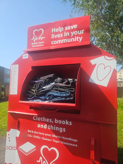 A photograph captures a red clothing donation bin from the British Heart Foundation situated outdoors, likely in Bristol, United Kingdom. The bin is filled with an assortment of clothing items. The bin’s design includes text that promotes clothing donation to “Help save lives in your community.” Adjacent text lists other acceptable donations such as “Clothes, books and things” and adds that shoes, handbags, CDs, DVDs, games, toys, and homewares are also welcome. A lower section outlines unacceptable donations like blankets, carpets, cushions, glass, metal, paper, pillows, quilts, rags, rubbish, rugs, and videos. The bin is set against a backdrop of green grass, a building, and a tree. The lighting suggests it is daytime.