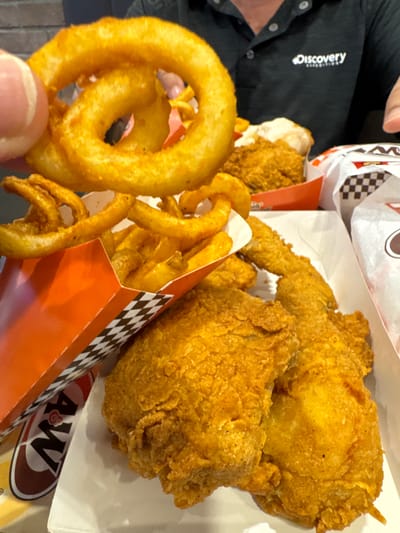 The image shows a close-up view of A&W fast food, including fried chicken pieces and curly fries, arranged in white and orange containers. A person's hand is visible in the upper left corner holding a curly fry. In the background, another person is wearing a black polo shirt with a "Discovery" logo. The setting appears to be indoors, likely a restaurant in Kuala Lumpur, Malaysia, given the presence of A&W.