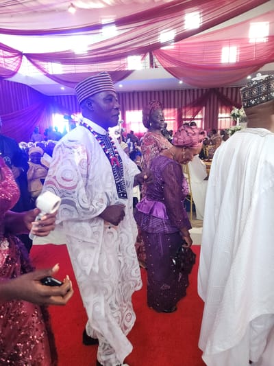 The image captures a gathering of people, likely at a social event such as a wedding, in Barden Yana, Nigeria. The scene is set indoors, with a red carpet covering the floor and draped fabric in shades of red and pink adorning the ceiling and walls, suggesting a festive atmosphere.

In the foreground, a man in traditional Nigerian attire, a white agbada with intricate lace detailing and a striped cap, is the central figure. He wears a colorful beaded necklace. To his left, a woman in a sequined red dress is partially visible, holding what appears to be a small white object in her right hand and a black object in her left. To the man's right, another man, also in white traditional attire and a cap, is seen from the back.

In the mid-ground, a woman in a purple lace outfit and a matching headscarf is looking down, with a beaded necklace visible. Behind her, other guests are milling about, some in traditional clothing. The background reveals more of the venue's decor and additional attendees, with glimpses of white chairs and floral arrangements. The lighting appears to be a mix of ambient and possibly colored stage lighting, casting a purple hue in some areas. The overall impression is one of a lively celebration.