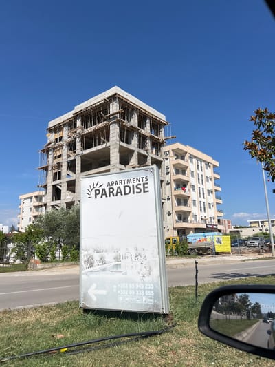 The image captures a street scene in Ulcinj, Montenegro, with a partially constructed building dominating the background. The building, made of concrete and wood scaffolding, is in the early stages of construction. To its right, a finished apartment building with beige and white exteriors stands, featuring multiple balconies and windows. In the foreground, a white billboard for "APARTMENTS PARADISE" is visible, with contact information and icons. A street runs in front of the buildings, with a road and grass verge. A yellow truck with colorful advertisements is parked behind the billboard. The sky is a clear, bright blue. The reflection in a car's side mirror shows a road with trees and buildings, further indicating an urban environment.