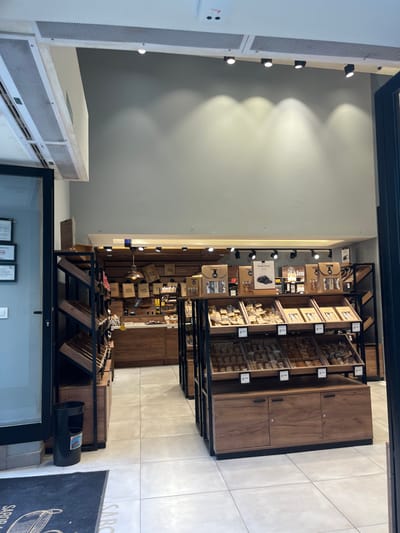 The image shows the interior of a shop located at coordinates 40.949, -4.122, which places it in the city of Segovia, Spain. The shop appears to be a gourmet food store, featuring a variety of packaged goods on shelves. The shelves are made of dark wood with black metal framing, and the products displayed on them are primarily boxed and packaged in brown paper. Several spotlights are mounted on the ceiling. There are no people present in the image, and the shop appears to be empty. The floor is tiled in a light gray color. An entrance doorway leads to a gray-tiled area with a black mat labeled "SARDI." A black trash can is visible at the bottom right of the image, and there is a partial view of a framed certificate on the bottom left. The scene suggests the shop is closed.