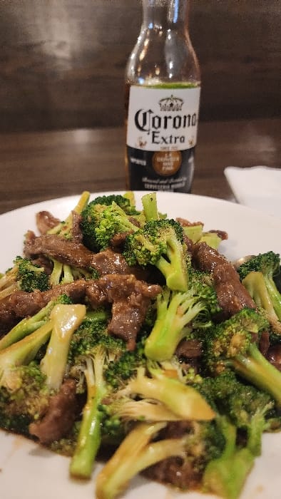 The scene depicts a meal and beverage served on a dark brown table surface in an indoor environment, located in Jackson, United States of America. In the foreground, a white, possibly oval-shaped plate contains a portion of beef and broccoli. The beef is presented as numerous irregularly shaped, dark brown pieces, coated in a glossy, brown sauce. Interspersed throughout the dish are vibrant green broccoli florets and stems, some with lighter green or yellowish bases, and small dark seasoning specks. Directly behind the plate, slightly to the right of the center, stands an opened, clear glass bottle of Corona Extra beer. The bottle features its characteristic white label with the "Corona Extra" logo, a crown emblem, and additional text in black and gold. The liquid level within the bottle is approximately one-third of its capacity, with a discernible green element at the liquid line, consistent with a lime wedge. The background is blurred and dark, indicating an interior setting, with a small, indistinct white object partially visible to the far right. No individuals, overt actions, or direct interactions are visible in the image.