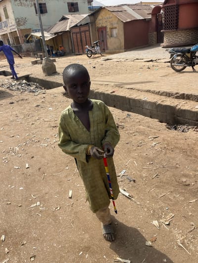 The image depicts a young, dark-skinned child standing centrally on an unpaved, dusty ground. The child wears a loose-fitting, long-sleeved olive-green garment, light-colored trousers, and dark sandals. Multiple gold-colored bangles are visible on their left wrist. In their hands, the child holds a vertically oriented object wrapped with multicolored fabric (red, yellow, blue, white). The child looks directly at the camera with a neutral expression.
To the child's left, a person in a blue outfit is partially visible, standing near a pile of debris in a ditch or gutter. Beyond them, a building with a light-colored facade and a partially visible sign that reads "BUILDING ZARIA" is present. Several other structures with corrugated metal roofs and varying wall finishes are visible in the background, along with a white motorcycle parked to the left.
To the child's right, a concrete curb or low wall runs horizontally across the mid-ground, separating the foreground's dirt path from a slightly elevated, possibly paved, area. More buildings with corrugated roofs and a parked motorcycle are visible further back. The ground throughout the scene is unpaved, covered in dirt, and scattered with debris including wood fragments and trash. Strong sunlight casts clear shadows. The specific location is identified as Zaria, Nigeria, based on the visible signboard.