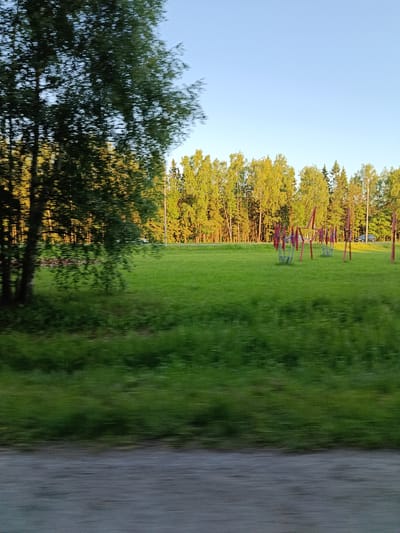 The image shows a motion-blurred view of a park-like area with a playground. The sun is setting, casting a warm glow on the trees in the background, which appear to be a mix of deciduous and coniferous. The sky is clear and blue. In the foreground, there is a grassy field with some playground equipment, primarily red metal structures that may be swings or climbing frames. The motion blur suggests the image was taken from a moving vehicle. There are no people clearly visible in the image due to the blur. The scene depicts an outdoor recreational space likely located in a suburban or park area.