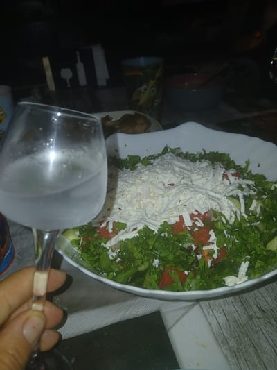 A close-up, slightly blurred, indoor shot shows a hand holding a wine glass filled with a clear liquid. In the background, a large white bowl overflows with a fresh salad composed of chopped lettuce, tomatoes, cucumbers, and generously topped with shredded white cheese. The setting appears to be a dining area, with hints of other tableware and decorative elements, including a black object with white silhouettes of a wine bottle and glass in the upper left. The scene is illuminated by artificial light, creating a subdued ambiance. The presence of the salad and wine glass suggests a meal is being enjoyed. The location is identified as Obrochishte, Bulgaria.