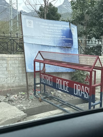 The image captures a scene in Dras, India, featuring a street-level view of a roadside barrier and a large informational sign. The barrier, painted red and blue, displays the words "DISTRICT POLICE DRAS" in white capital letters. A small red stop sign with a skull icon is affixed to the barrier. Behind the barrier, a large blue and white sign is partially visible, containing text in English and what appears to be Urdu. The English text includes "U.T OF LADAKH LEGAL SERVICES AUTHORITY FREE LEGAL SERVICES (LEGAL AID)" and lists categories of individuals eligible for free legal services. The environment appears somewhat disheveled, with debris scattered around the base of the sign and barrier. In the background, a mountainous landscape is visible, suggesting the mountainous terrain of the region.