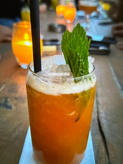 A clear glass, containing an orange-hued beverage with a frothy white head and ice cubes, is positioned on a light-colored coaster or napkin. A dark-colored straw is inserted into the drink. A cluster of fresh green mint leaves serves as a garnish, rising from the ice. A faint, light grey watermark reading "TEN MODERN ASIAN" is visible on the ice. The immediate surface beneath the glass is a wooden table with visible grain. The background is out of focus, revealing an amber-colored glass or candle holder with a lit flame to the left, and additional blurred elements consistent with a restaurant or bar setting. No individuals are discernible. This scene is located in Senokos, Bulgaria, likely within the establishment identified as Ten Modern Asian.