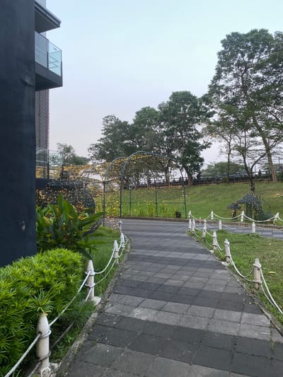 An outdoor pathway in a park area is depicted. The pathway is paved with dark gray and black square tiles, with a white rope and post barrier running alongside. To the left of the pathway, a dark gray building with modern architecture, featuring balconies with glass railings and horizontal louvers, is visible. In front of the building and along the pathway, there is lush green foliage. Beyond the pathway, an archway decorated with numerous small, warm-toned lights is visible. Further into the park, more trees and a grassy slope can be seen, with a small, gazebo-like structure also adorned with lights. The sky is overcast and pale. The scene appears to be in a public park in East Jakarta, Indonesia, possibly at dusk or early morning given the artificial lighting. No people are clearly visible or identifiable in the image.