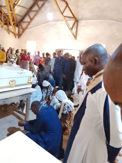 The image shows a funeral ceremony taking place in a simple building with a wooden roof. The room is sparsely decorated with white walls and exposed wooden beams. A white casket is displayed on a wooden table in the foreground. There are several people seated on the floor in front of the casket, some kneeling and some standing, with their heads bowed in prayer or mourning. Other people are standing in the background, observing the scene. The people in the image are all dressed in various clothing, including traditional attire. The general atmosphere is somber and subdued. The location of the image is not possible to determine based on the provided coordinates, as these coordinates are located in a forested area in Nigeria, not an urban center.