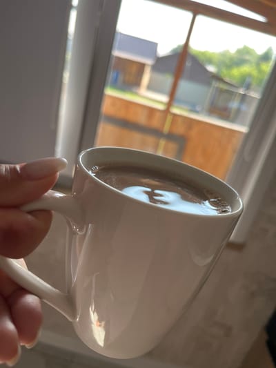 A hand with light-colored nails holds a white mug filled with a dark beverage, possibly hot chocolate or coffee, near a window. The mug is white ceramic and has a handle. The beverage has a foamy surface with some bubbles. Outside the window, a wooden fence is visible, along with houses and green trees. The scene appears to be indoors, with the light suggesting it is daytime. The image is taken at a close-up perspective, focusing on the mug and the hand. No specific location in Novosibirsk, Russia, can be identified from the visual information provided.