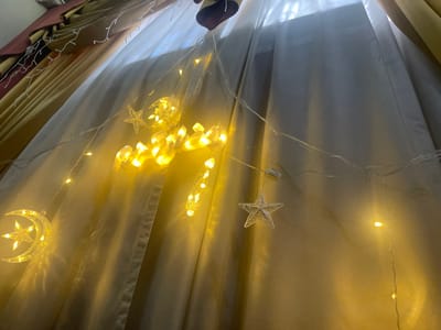 An indoor scene within the Sarouja Municipality, Syria, depicts illuminated decorative string lights arranged against a backdrop of drawn window curtains. The primary subject is a string of warm yellow LED lights with transparent plastic ornaments, including a crescent moon with an embedded star on the lower left, an additional star, and a central arrangement of stylized forms resembling Arabic script. Smaller, non-illuminated star-shaped ornaments are also present along the clear wiring. These lights are positioned in front of a translucent white or off-white curtain that covers a window. To the upper left, a heavier, pleated curtain in a yellow-gold color is partially visible behind the sheer layer. No individuals or specific actions beyond the illumination of the lights are depicted.