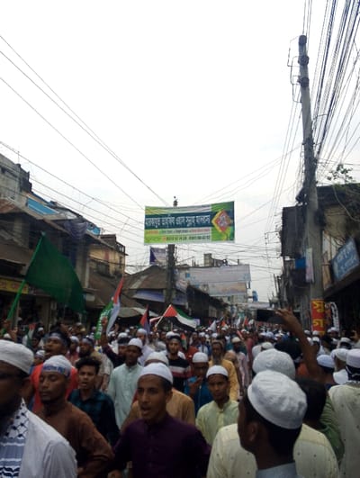 The image shows a large crowd of people gathered in the street in Bhola, Bangladesh, at the coordinates 22.686, 90.644. They are all men, and most are wearing white skullcaps. Some are holding up cellphones and waving flags. Many of the men are wearing long white shirts. There is a large banner hanging above the crowd that is written in Bengali. In the background there are buildings lining the street. The sky is cloudy and there are many power lines strung across the sky. The crowd appears to be a religious procession.