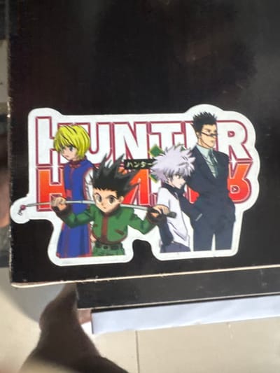 The image presents a rectangular sticker affixed to a dark, flat surface, possibly a box or shelf, located in Ipetumodu, Nigeria. The sticker depicts four anime characters from "Hunter x Hunter" standing in a line, with the series title "HUNTER HxH" displayed in large red and white lettering, accompanied by smaller Japanese text. From left to right, the characters are: a blond-haired individual in a blue tunic; a dark-haired character in a green outfit, holding a fishing rod horizontally across his shoulders; a white-haired character in a white shirt; and a dark-haired character wearing glasses and a dark suit. The dark surface shows minor surface wear. Below the surface, light-colored floor tiles are visible, and a blurred, dark-skinned human foot is partially present in the lower left foreground.