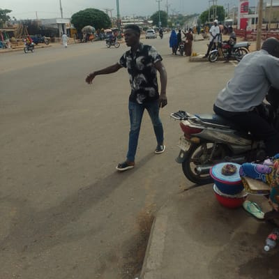 The image captures a bustling street scene in Tumfure, Nigeria, under an overcast sky, suggesting a cloudy or potentially rainy day. The primary focus is a young man in the foreground, wearing a black t-shirt with a white pattern and blue jeans, walking across the asphalt road. He is looking to his right with an open-mouthed expression, possibly calling out or reacting to something. He wears black and white sandals.
Behind him, several motorcycles are visible. One is ridden by a person wearing a grey shirt and dark pants, another by a person in a red shirt and helmet. Further in the background, there's a variety of activity: people walking, several motorcycles, and some wares displayed on the side of the road. In the distance, buildings and trees line the street, with overhead power lines indicating an urban or semi-urban setting. Some construction work with scaffolding and building materials is also apparent on the left side of the image.
On the right side of the foreground, a motorcycle is parked, with colorful woven baskets stacked on bowls next to it. A light blue flip-flop lies on the ground near the motorcycle. A plastic bottle with a red label, likely a beverage, is also visible on the ground. The overall impression is one of everyday life and commerce taking place on a busy street. There is a sign visible in the far background with some text, but it is too blurry to read.