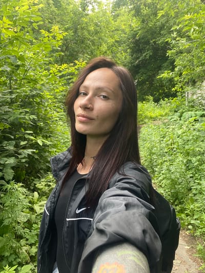 The image depicts a woman with long, dark brown hair and fair skin, wearing a black Nike jacket and a black t-shirt. She has a nose ring and a small stud below her lower lip. The background is lush green foliage, suggesting an outdoor, natural setting, possibly a park or a wooded area within the city of Oktyabrsky, Russia. The lighting indicates it is daytime. The woman is holding a device, likely a phone, to take a selfie. The scene is composed from a chest-up perspective, with the woman's face as the focal point.