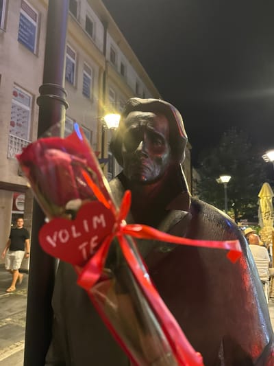 The image shows a close-up of a statue at night in the city of Tuzla, Bosnia and Herzegovina. A red rose with a red heart-shaped tag that says "VOLIM TE" (I love you) is placed in front of the statue. The statue appears to be made of bronze and depicts a man with a serious expression. In the background, there are buildings with illuminated windows, streetlights, and some people walking or sitting at outdoor tables. The overall lighting suggests it is evening or night.