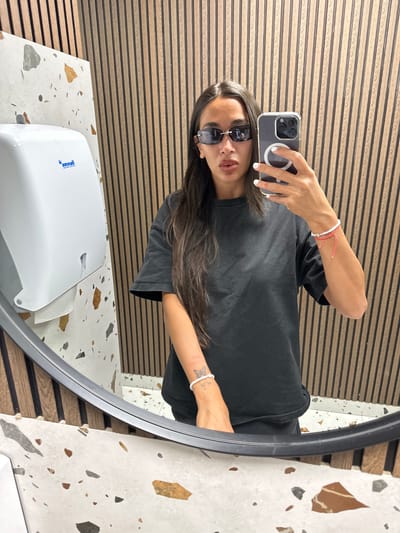 A woman takes a mirror selfie in a modern restroom. She is wearing dark sunglasses, a black t-shirt, and has long, dark hair. She is holding a smartphone in her right hand, which is pointed towards the mirror. On her left wrist, she is wearing a white bracelet with a butterfly charm and a pink string bracelet. The restroom features a white paper towel dispenser on the left wall, with a speckled white and brown tile backsplash. The background wall is made of vertical wooden slats. The floor is also covered in the speckled tile. The location is not identifiable as Pernik, Bulgaria.