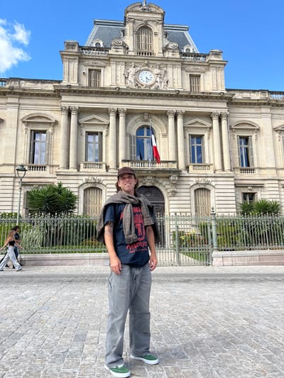 A man stands in front of Montpellier City Hall in Montpellier, France. The man wears a brown hat, a navy t-shirt with red text, gray trousers, and green sneakers. A brown jacket is draped over his shoulders. The city hall, a multi-storied stone building with ornate architectural details, including columns and a clock tower, forms the backdrop. A French flag hangs from the building. A wrought iron fence runs along the front of the building with foliage visible behind it. Two figures, one in white pants and the other in a black top, are partially visible in the lower left corner. The ground is paved with light-colored stone bricks. The sky is blue with some clouds.