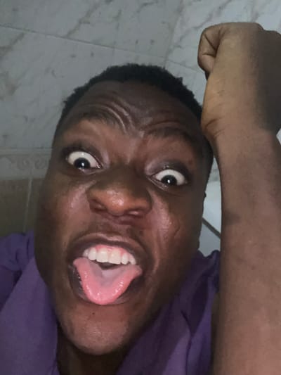 A close-up, slightly angled, indoor photograph shows a dark-skinned person with their tongue sticking out. Their eyes are wide. They are wearing a purple garment. The person has short, dark hair. A clenched fist is visible above their head. The background appears to be a tiled wall. No specific details point to Ikorodu, Nigeria.
