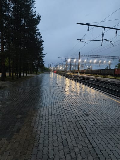 The image depicts a train station platform in what appears to be Magdagachi, Russia. It is twilight or nighttime, with an overcast sky. The platform is constructed of interlocking pavement stones. The right side of the platform is wet. Train tracks run along the right side of the platform, parallel to the length of the image. Railroad cars are visible in the distance. Overhead power lines and support structures are strung above the train tracks. A row of trees is along the left side of the platform. Streetlights illuminate the train tracks and surrounding area.