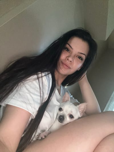This portrait-oriented image features a young woman and a small white dog in an indoor setting, presumably in Sliven, Bulgaria. The woman, with long, dark hair flowing over her shoulders, is seen from a low angle, looking directly at the camera with a relaxed, gentle smile. Her face shows natural skin texture, possibly freckles, and she wears a simple white t-shirt and a thin necklace. Her right arm is bent, with her hand near her head, and her bare legs are visible in the upper right of the frame, suggesting she is lounging or reclining.
Nestled closely against her chest is a small, white dog, resembling a Chihuahua, with large ears, dark eyes, and a dark nose. The dog is also looking towards the camera, appearing calm and comfortable. The background is indistinct, showing light-colored walls or a ceiling, typical of an interior space. Soft, natural light illuminates the scene, suggesting it is daytime. The overall impression is one of affectionate companionship and relaxation. No text is visible in the image.