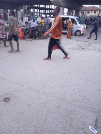A busy street scene unfolds under an elevated highway structure in Port Harcourt, Nigeria. In the foreground, a man in an orange and black tracksuit walks past, his form blurred by motion. His feet are adorned with bright orange flip-flops. To his left, a man in a light brown shirt and dark shorts walks away from the camera. Further back, a cluster of people and vehicles indicates commercial activity. A silver minivan is parked, with individuals entering and exiting. Several motorcycles and a red cargo bag are visible near a group of seated individuals. The environment appears to be a paved street with some debris scattered on the ground. The overall impression is one of a dynamic urban environment with constant movement and pedestrian and vehicular traffic.