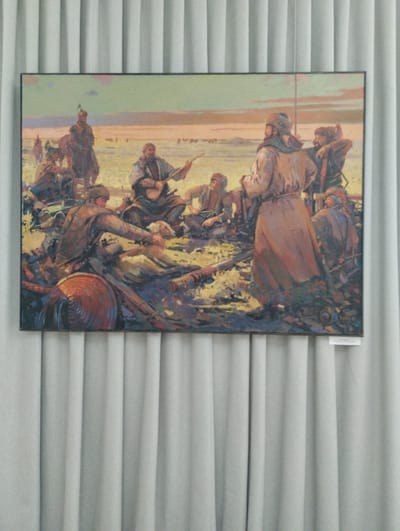 The image shows a painting depicting a group of people in what appears to be a historical or cultural setting, likely in Kazakhstan, given the location context. The painting features several individuals, some on horseback and others gathered around a fire or a campsite. The figures are dressed in traditional clothing, including fur hats and long robes, suggesting a nomadic or historical theme. One person in the center is playing a stringed instrument, perhaps a dombra, indicating a musical or social gathering. Other individuals are engaged in various activities, such as tending to a fire or interacting with each other.
The scene is set in an open, arid landscape with a muted color palette, suggesting either dawn or dusk. The sky is painted with soft hues of orange and yellow, hinting at a warm atmosphere. The overall mood of the painting seems to be one of communal gathering and storytelling, with a sense of historical significance. There are also some objects visible, including what might be a saddle and other equipment, further reinforcing the historical and nomadic theme. There is a small white label attached to the bottom right of the frame, but the text is not legible. The painting is framed and hung against a backdrop of grey vertical blinds.