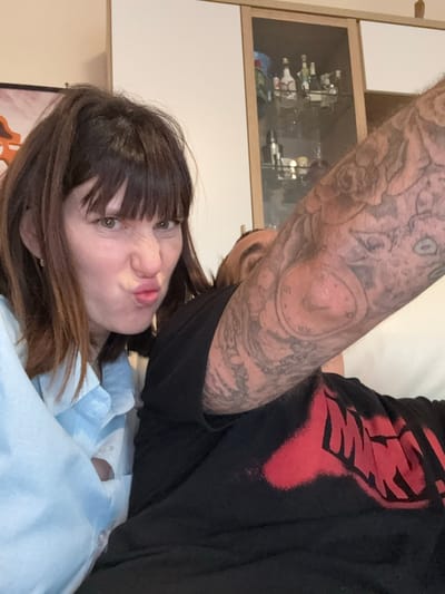 A close-up selfie captures a woman with brown hair and bangs, making a pouting facial expression. She is wearing a light blue shirt. Over her shoulder is a man with a heavily tattooed arm. The man wears a black shirt with a red design. Behind them is a cabinet with glass doors containing bottles. The background features white walls and wooden elements. The location in the city of Aprilia, Italy cannot be confirmed from the image.