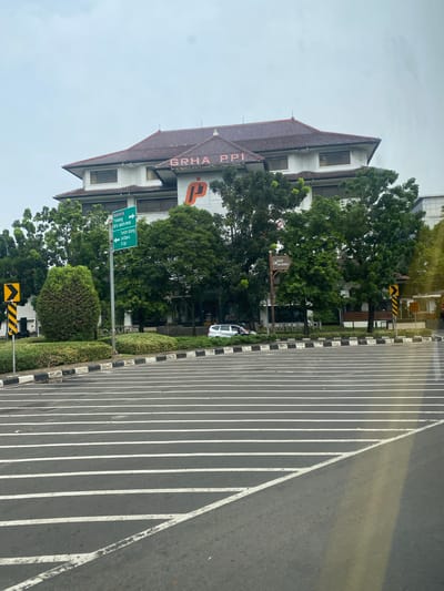 The image shows a building with a red tiled roof, white walls, and dark windows. The building has a sign that reads "GRHA PPI" in large pink letters, with a smaller orange logo to the right. In front of the building, there are several green trees and bushes. A traffic sign with green arrows indicates directions to "Batavia", "Tomang", "Tol Jakarta-Merak", "Tanah Abang", "Jatibaru", and "Slipi". Another sign, a brown arrow, points towards "MUSEUM BANK INDONESIA". The foreground shows a road with white painted lines, suggesting a pedestrian crossing or a lane marking. A car and a motorcycle are visible in the background. The sky is overcast. The building is identified as the "GRHA PPI" located in Central Jakarta, Indonesia, which is likely the office of the Securities Investor Protection Fund.