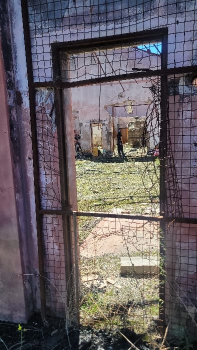 The image captures a view through a heavily rusted and distorted metal framework, covered by a torn wire mesh, likely an entrance or window opening within a derelict structure in Zárate, Argentina.

The immediate foreground features the dark, angular metal frame and its square-grid wire mesh, which is significantly broken and twisted, particularly on the right side. Dry vegetation and darkened ground are visible at the very bottom.

Beyond this barrier, an outdoor area is revealed under bright daylight. The ground consists of uneven terrain with patches of green grass, interspersed with dirt, debris, and what appears to be a concrete block lying flat towards the lower right.

In the mid-ground, a dilapidated building wall, painted a faded pink or peach color, shows extensive signs of decay, including peeling paint, exposed masonry, and structural damage. Several irregular openings or doorways are visible in this wall, leading to interior spaces or further sections of the ruin. Above the visible wall sections, a clear blue sky is apparent, suggesting a missing or collapsed roof structure.

Two individuals are observable in the background. On the right, one person stands upright, dressed in dark clothing, with their back partially turned to the viewer, facing towards the left. Further to the left, near another opening in the wall, a second individual is partially visible, appearing to be in a crouched or bent-over position, also wearing dark attire. Debris, including a yellow object, is scattered on the ground near this individual.