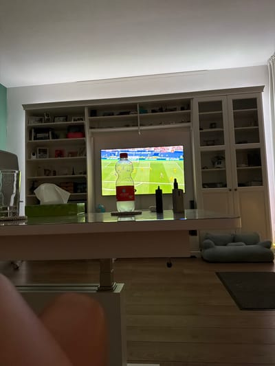 The image depicts a living room scene with a television playing a soccer match. In the foreground, a glass of water, a box of tissues, and a bottle of "water" are placed on a glass-top coffee table. Two vape pens are also visible on the table. Behind the coffee table, a large bookshelf unit is filled with various items, including books, picture frames, and decorative objects. The television screen shows a soccer game with players on a green field, and the text "WORLD CUP 2022" is partially visible. To the right of the bookshelf, a light blue pet bed is placed on the wooden floor. The overall environment suggests a casual indoor setting. There is no indication of the specific location within Helmond, Netherlands, in the image.