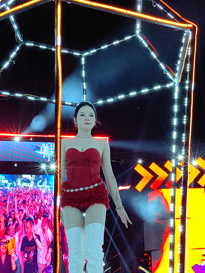 An eye-level, full shot photograph shows a young woman in a red bustier top and red shorts, along with white boots, standing on a stage with a light display. This appears to be a performance in Bai Chay Ward, Vietnam. A large screen on the left shows the audience, who are visible in the crowd. The stage has orange and white lights. The woman is fair-skinned with dark hair pulled back, wearing a necklace and bracelet. She has a tattoo on her right forearm. She appears to be performing for the crowd.